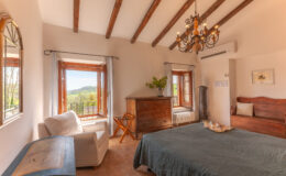Cozy bedroom with wooden beams, featuring a comfortable bed, vintage dresser, and large windows offering scenic views. The room is decorated with a chandelier and a potted plant, creating a warm and inviting atmosphere.