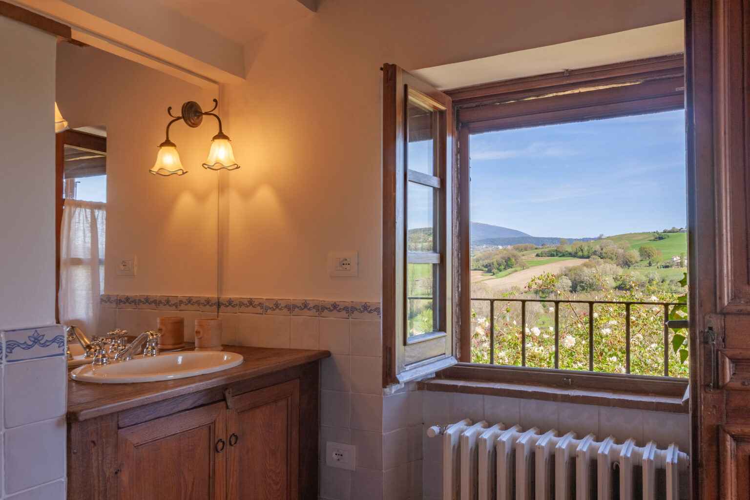 Bright and inviting bathroom featuring a wooden vanity and sink, with a large window offering a picturesque view of rolling green hills and blue sky. Natural light enhances the warm tones of the decor. | Only on SopranoVillas