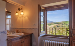 Bright and inviting bathroom featuring a wooden vanity and sink, with a large window offering a picturesque view of rolling green hills and blue sky. Natural light enhances the warm tones of the decor.