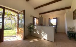 Bright and airy kitchen interior with wooden beams, featuring an open door leading to a garden view, modern white cabinetry, and natural light illuminating the space.