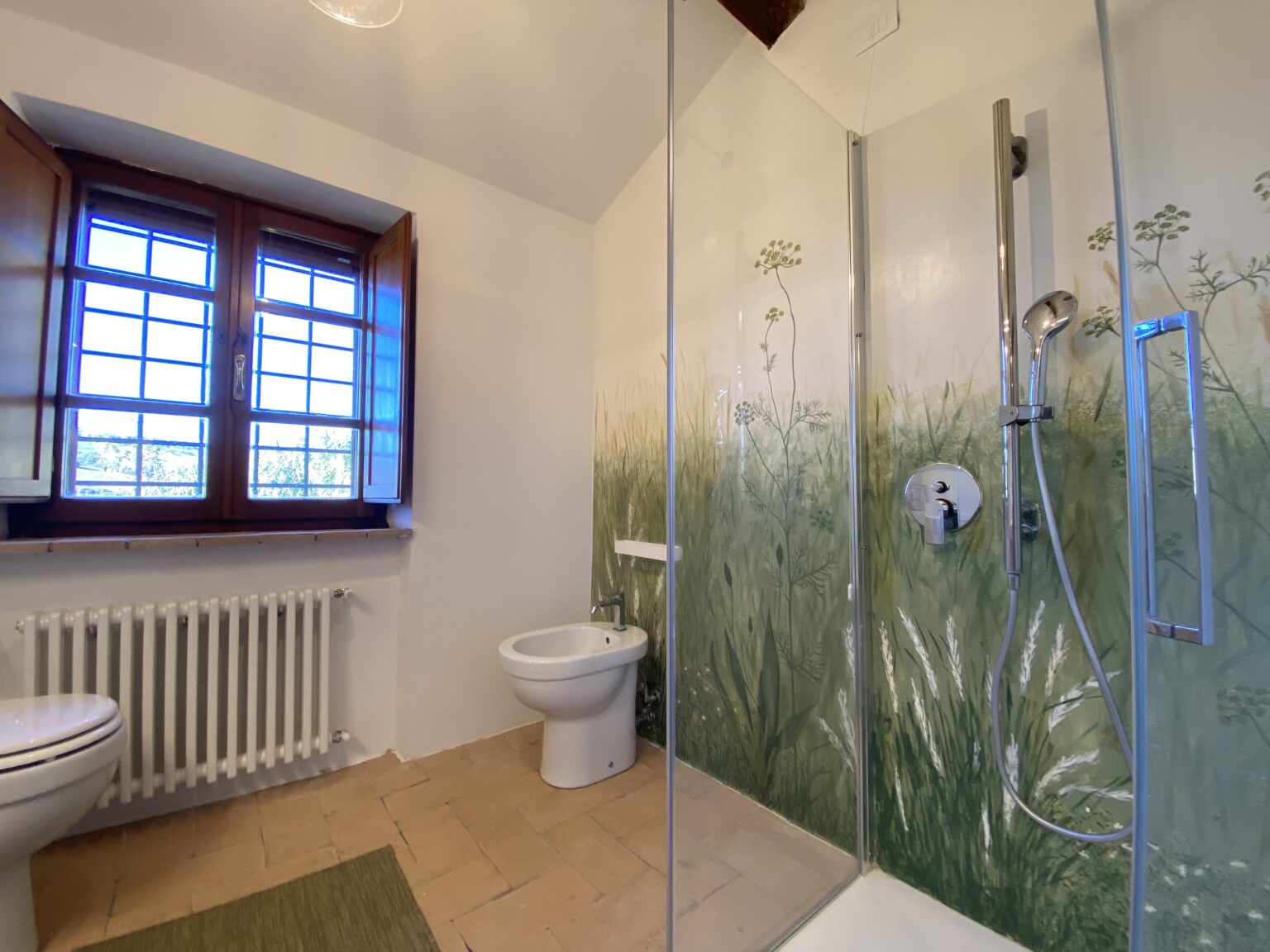 Modern bathroom featuring a glass shower, toilet, and a decorative nature-themed mural. Natural light streams in through wooden windows, enhancing the serene atmosphere. | Only on SopranoVillas