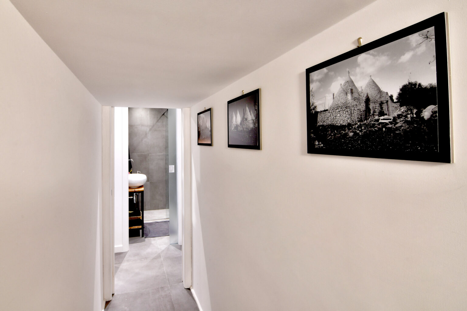 Narrow hallway featuring white walls and black and white framed photographs, leading to a modern bathroom with gray accents. | Only on SopranoVillas