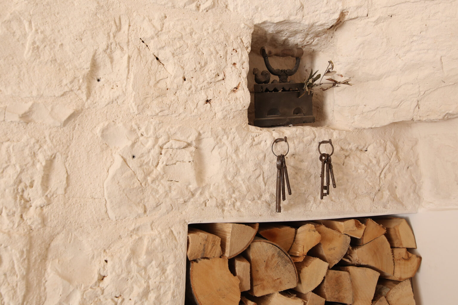 Rustic wall decor featuring an antique key holder with vintage keys hanging beside a wood storage area filled with neatly stacked firewood, set against a textured white stone wall. | Only on SopranoVillas