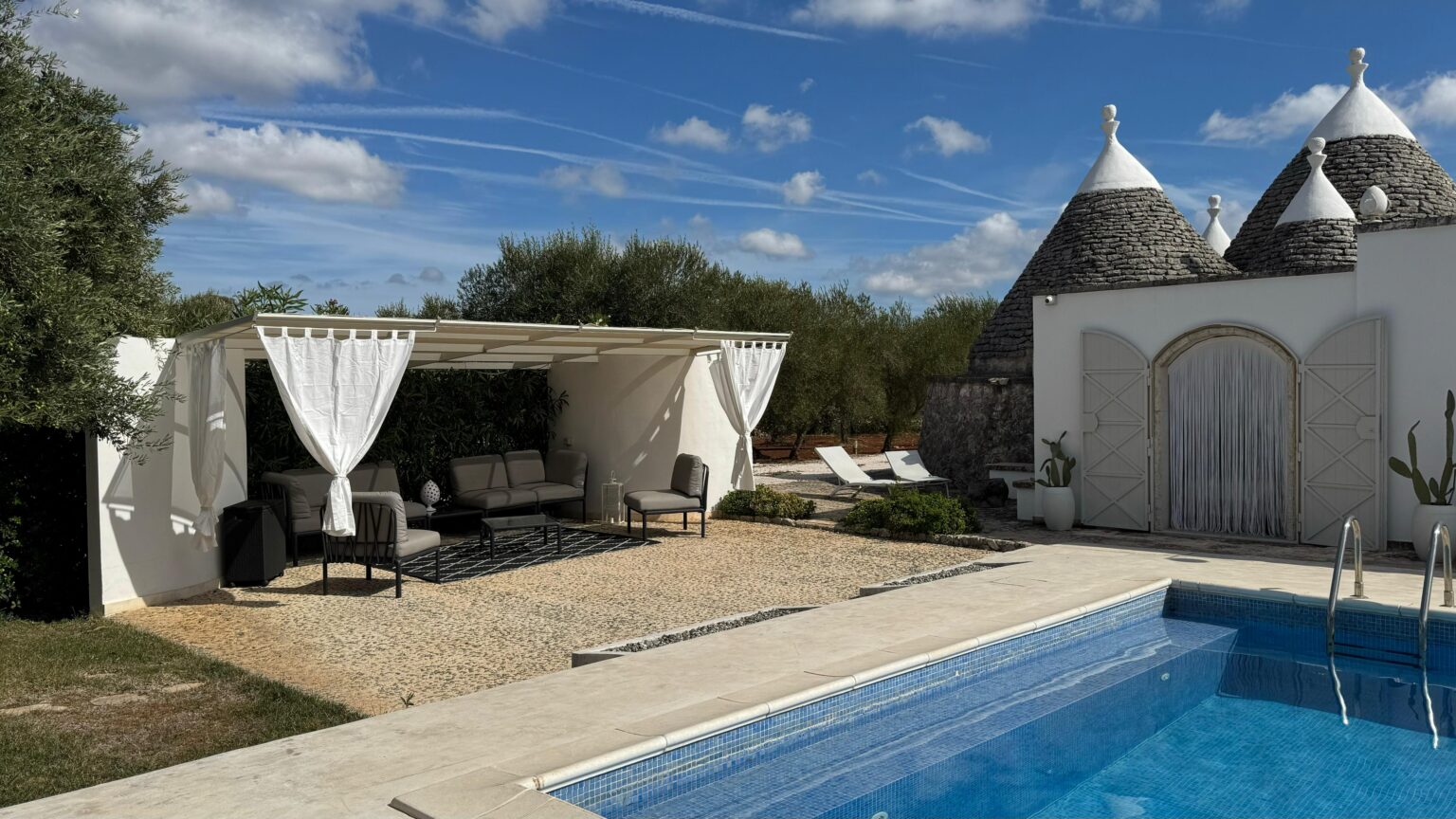 Tranquil outdoor living space featuring a pool, a shaded seating area with curtains, and traditional trullo architecture under a bright blue sky. Ideal for relaxation and leisure. | Only on SopranoVillas