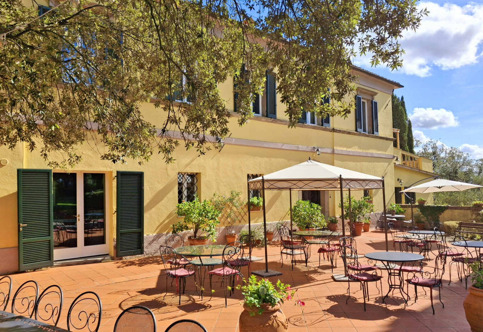Charming outdoor café terrace with tables and chairs under a sunny sky, surrounded by greenery and a yellow building, perfect for dining al fresco. | Only on SopranoVillas