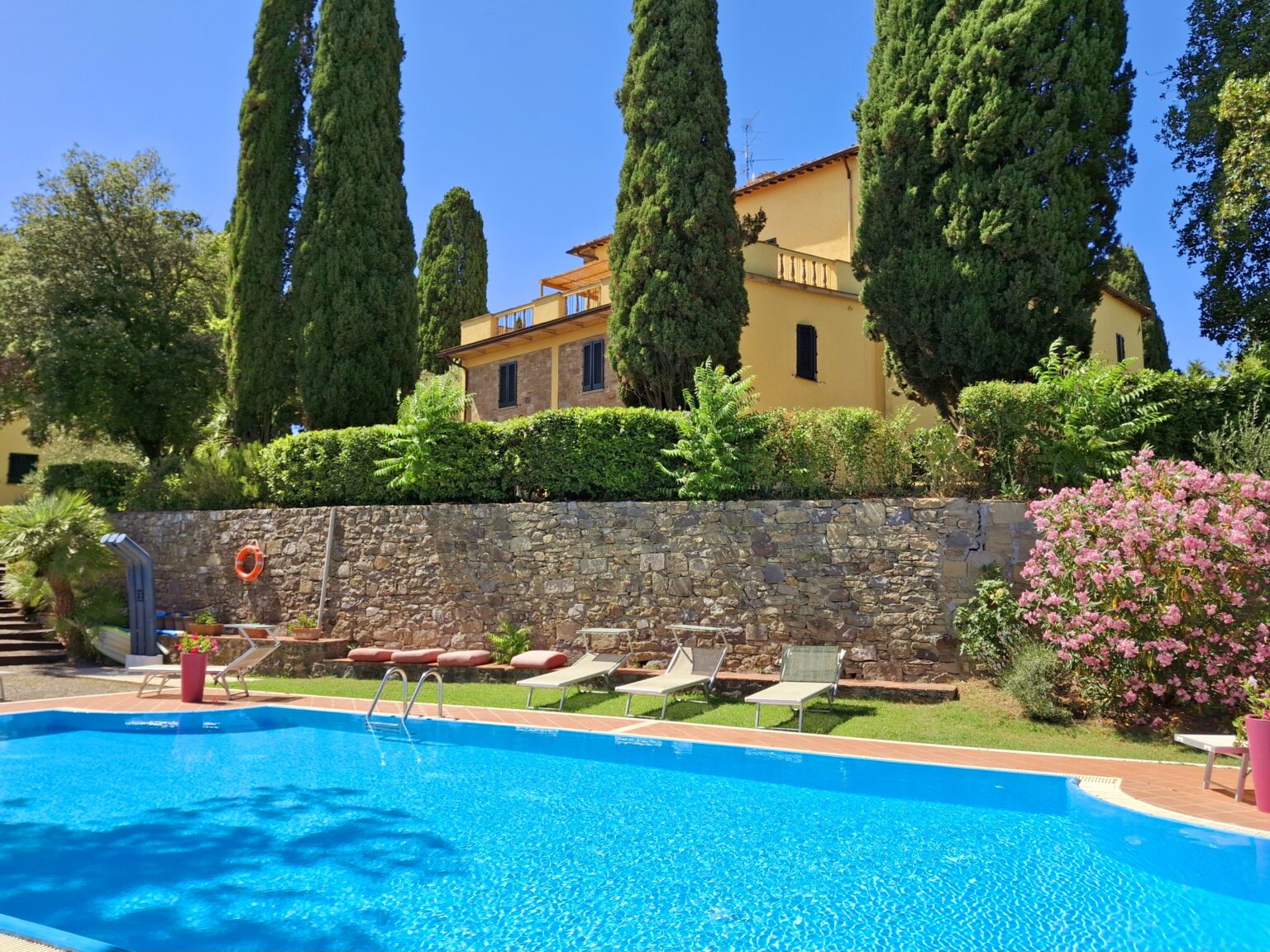 Scenic view of a luxurious villa with a swimming pool surrounded by lush greenery and colorful flowers, featuring comfortable lounge chairs and tall cypress trees under a clear blue sky. | Only on SopranoVillas