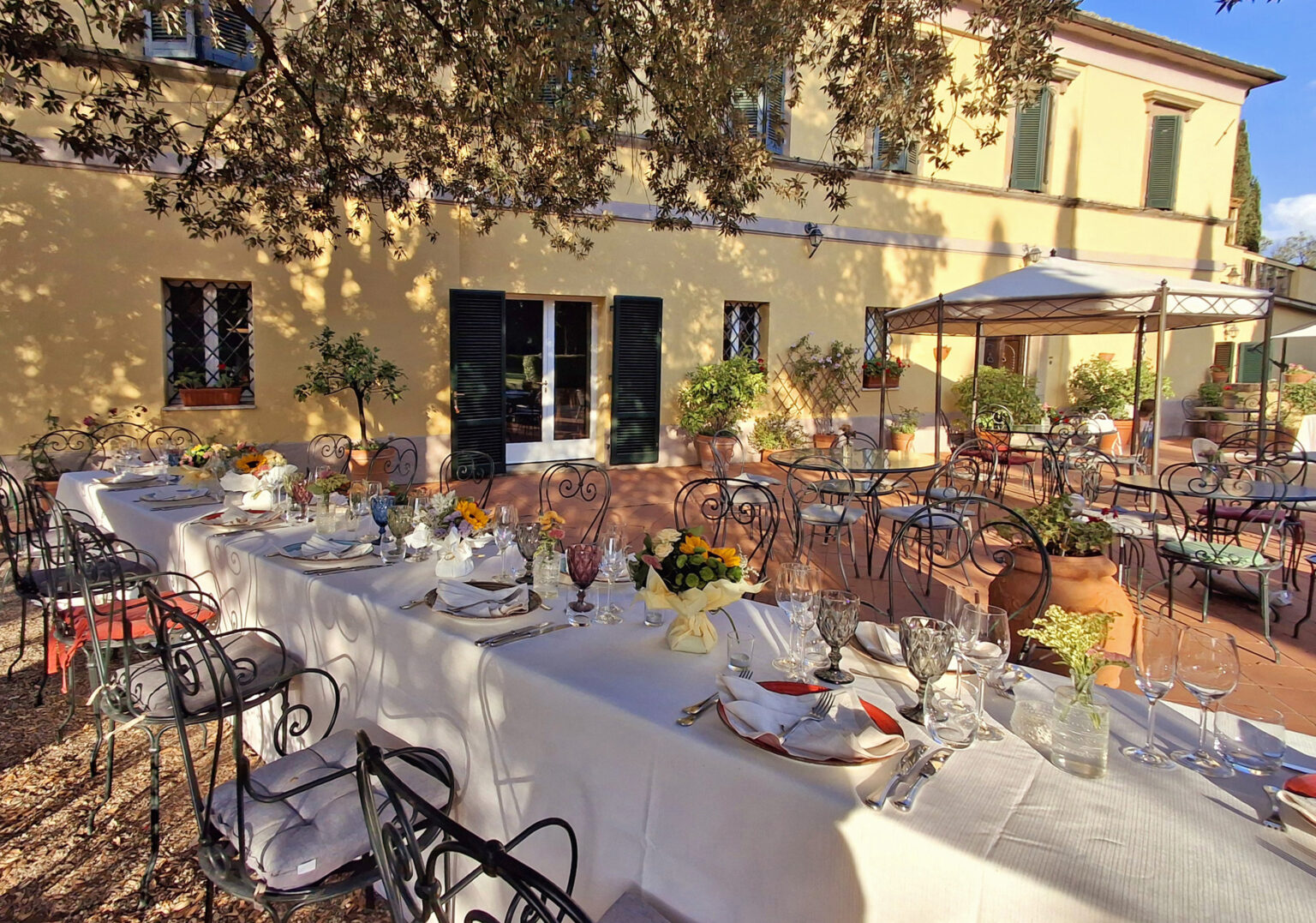 Outdoor dining setup at a charming restaurant with elegantly arranged tables, white tablecloths, and decorative floral centerpieces, surrounded by greenery and a sunny atmosphere. | Only on SopranoVillas