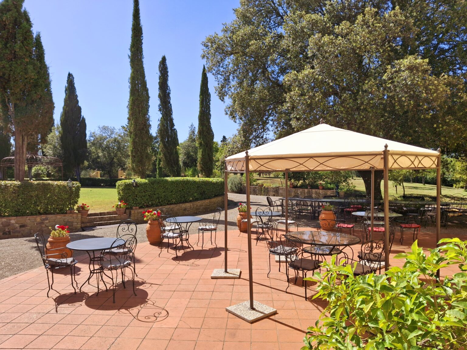 Outdoor dining area featuring metal tables and chairs under a large canopy, surrounded by lush greenery and tall cypress trees in a sunlit garden setting. Ideal for events or casual gatherings. | Only on SopranoVillas
