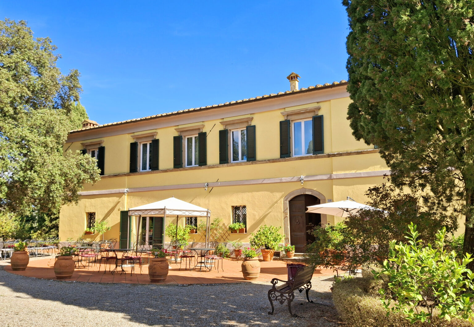 Charming yellow villa with green shutters, featuring a spacious outdoor terrace with umbrellas and potted plants, set against a clear blue sky. Ideal for vacation rentals or outdoor gatherings. | Only on SopranoVillas
