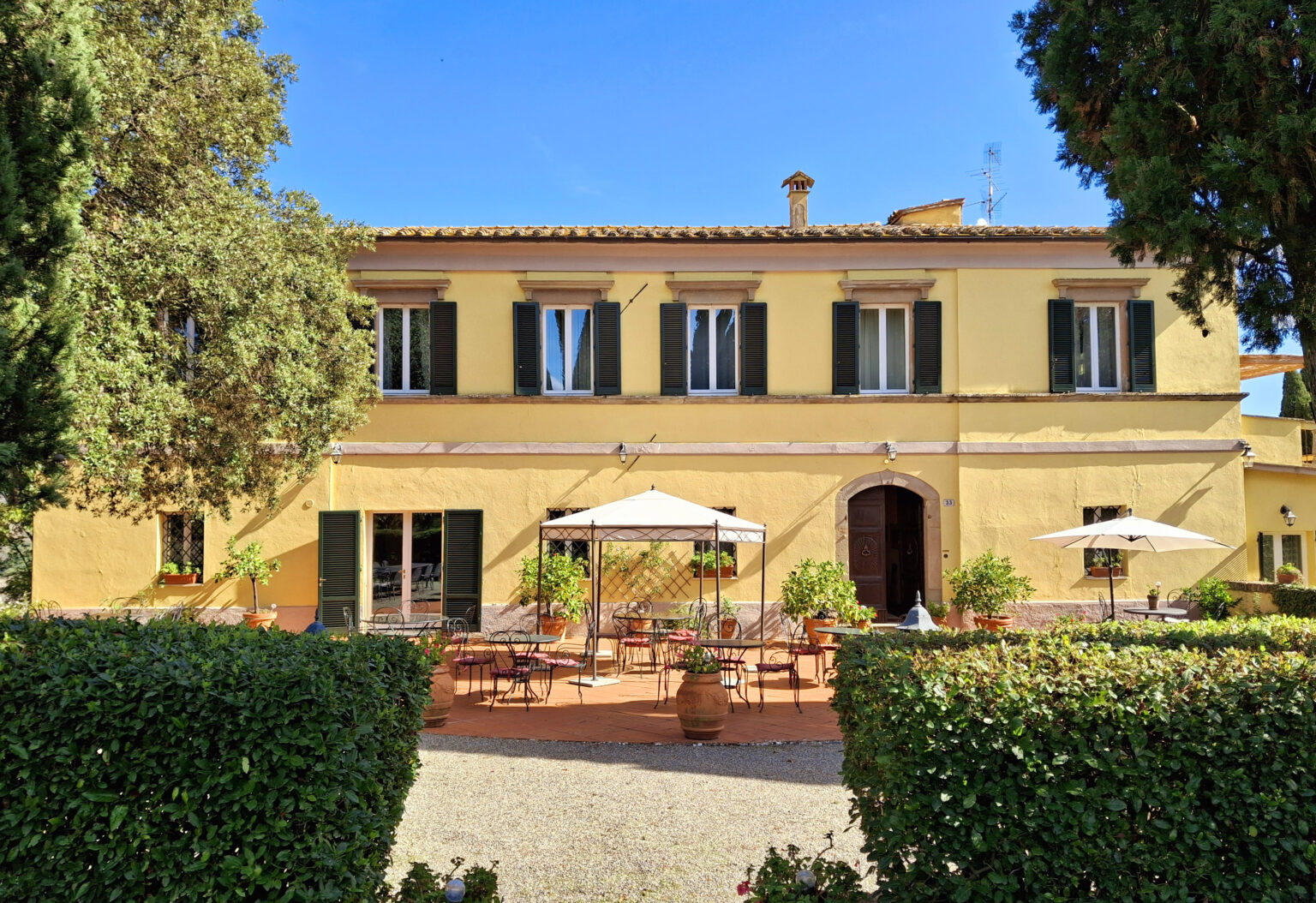 Charming yellow villa with outdoor seating and umbrellas, surrounded by greenery under a clear blue sky. Ideal for relaxation and dining al fresco. | Only on SopranoVillas