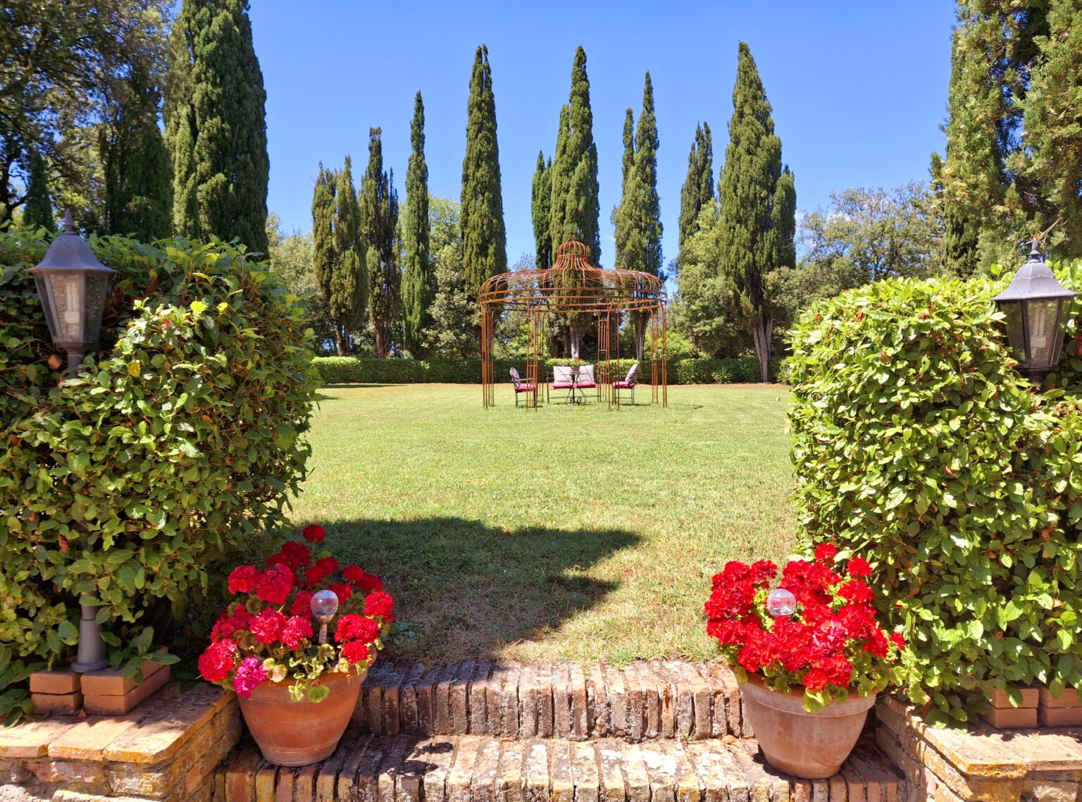 Lush garden with vibrant red flowers in pots, framed by neatly trimmed hedges, leading to a grassy area with tall cypress trees and a wooden gazebo in the background, under a clear blue sky. | Only on SopranoVillas