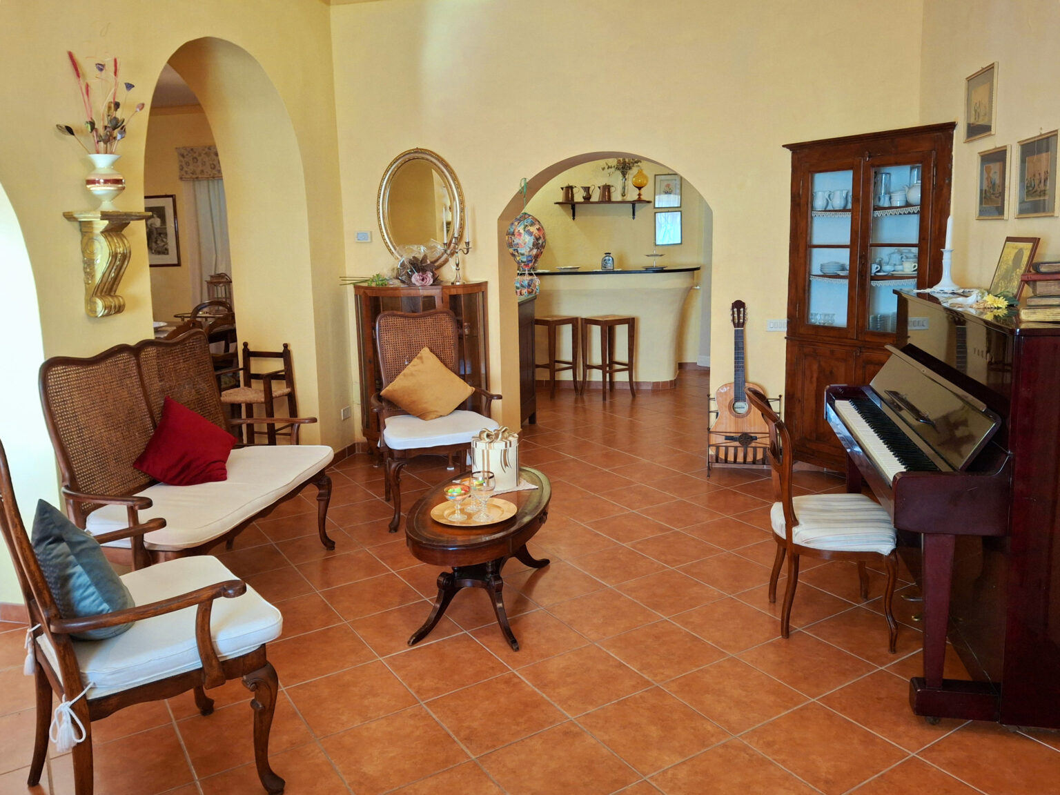 Cozy living room featuring vintage furniture, a grand piano, and warm terracotta flooring, with an inviting archway leading to a bar area. | Only on SopranoVillas