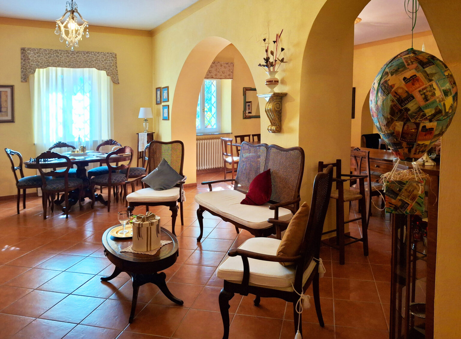 Cozy living room interior featuring vintage furniture, a round dining table, and warm yellow walls. The space is illuminated by a chandelier and decorated with a unique hot air balloon ornament. Comfortable seating arrangements include plush cushions on wooden chairs, creating an inviting atmosphere. | Only on SopranoVillas