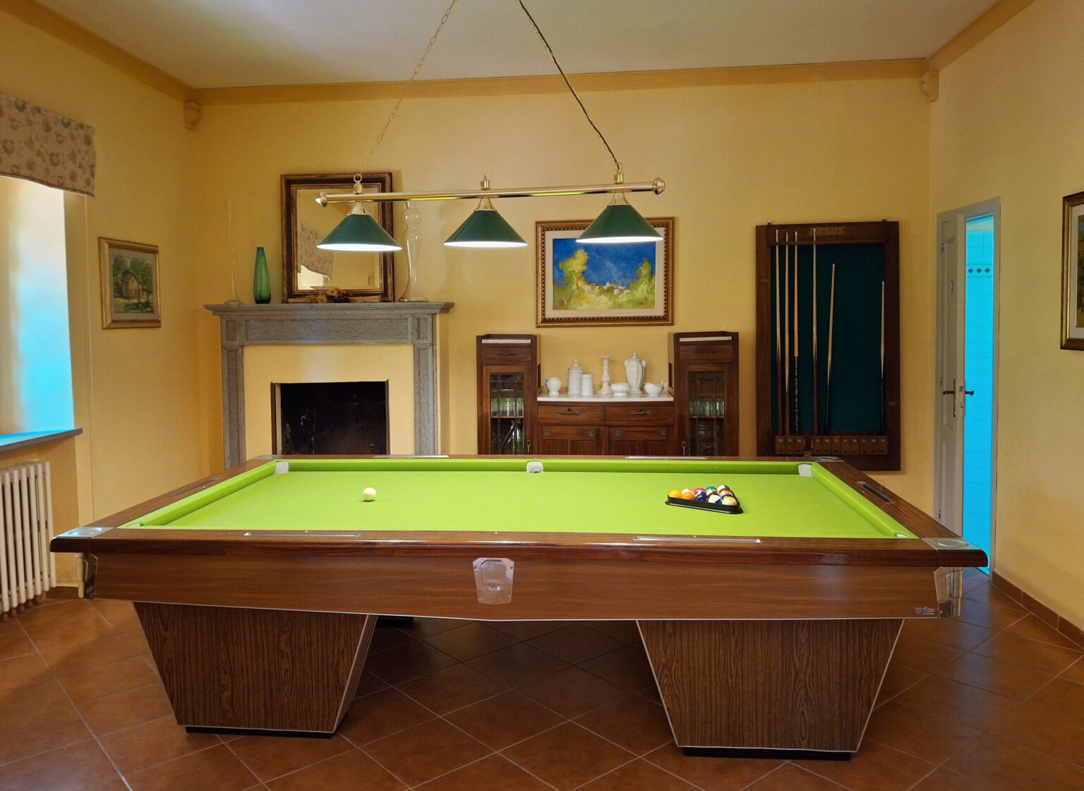 Interior of a game room featuring a green billiards table, wooden furniture, and decorative paintings on yellow walls, with ambient lighting from overhead lamps. | Only on SopranoVillas