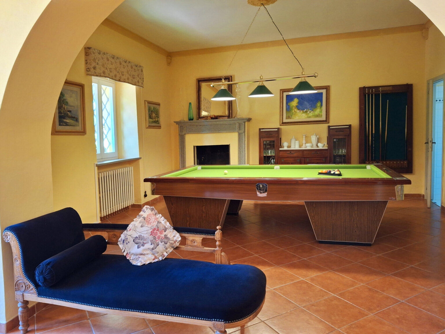 Elegant game room featuring a green billiard table, vintage decor, and a comfortable blue chaise lounge with floral accents. The yellow walls and natural light create a warm atmosphere, complemented by artwork and a stylish fireplace. Perfect for leisure and entertainment. | Only on SopranoVillas