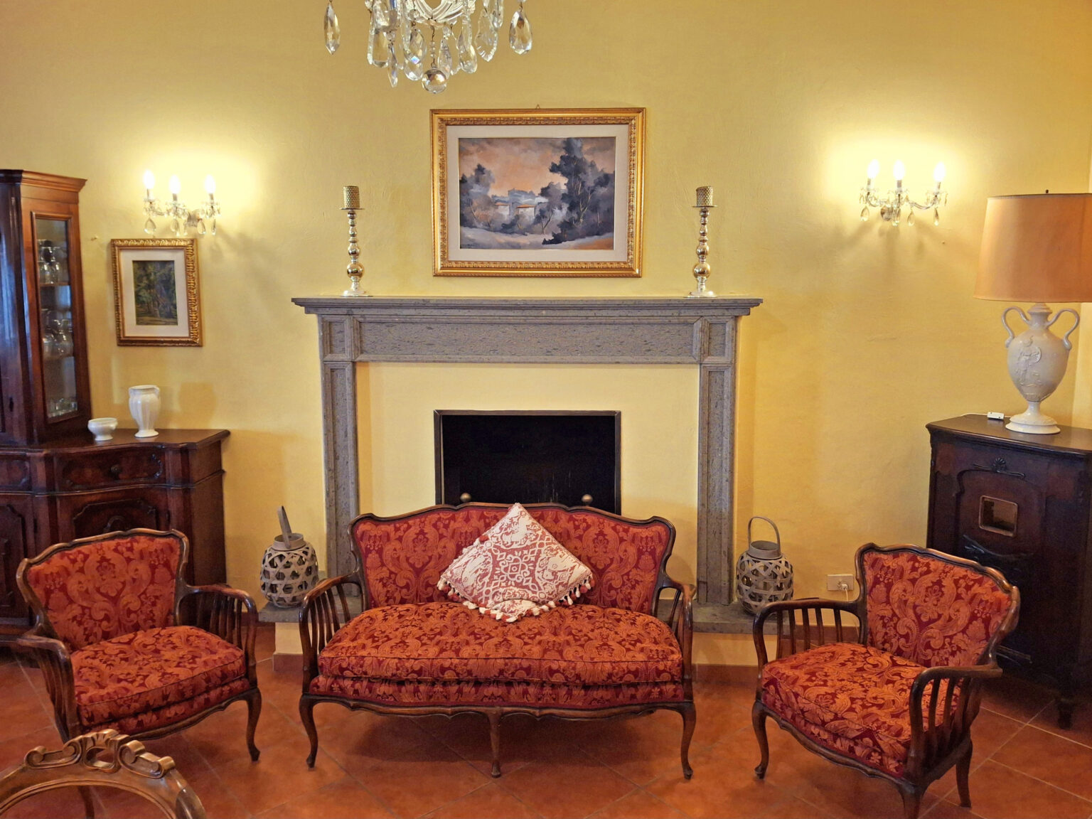 Cozy living room with vintage furniture, featuring an ornate fireplace, a classic painting above the mantel, and elegant wall sconces. The seating includes a floral-patterned sofa and two matching chairs, creating a warm and inviting atmosphere. | Only on SopranoVillas