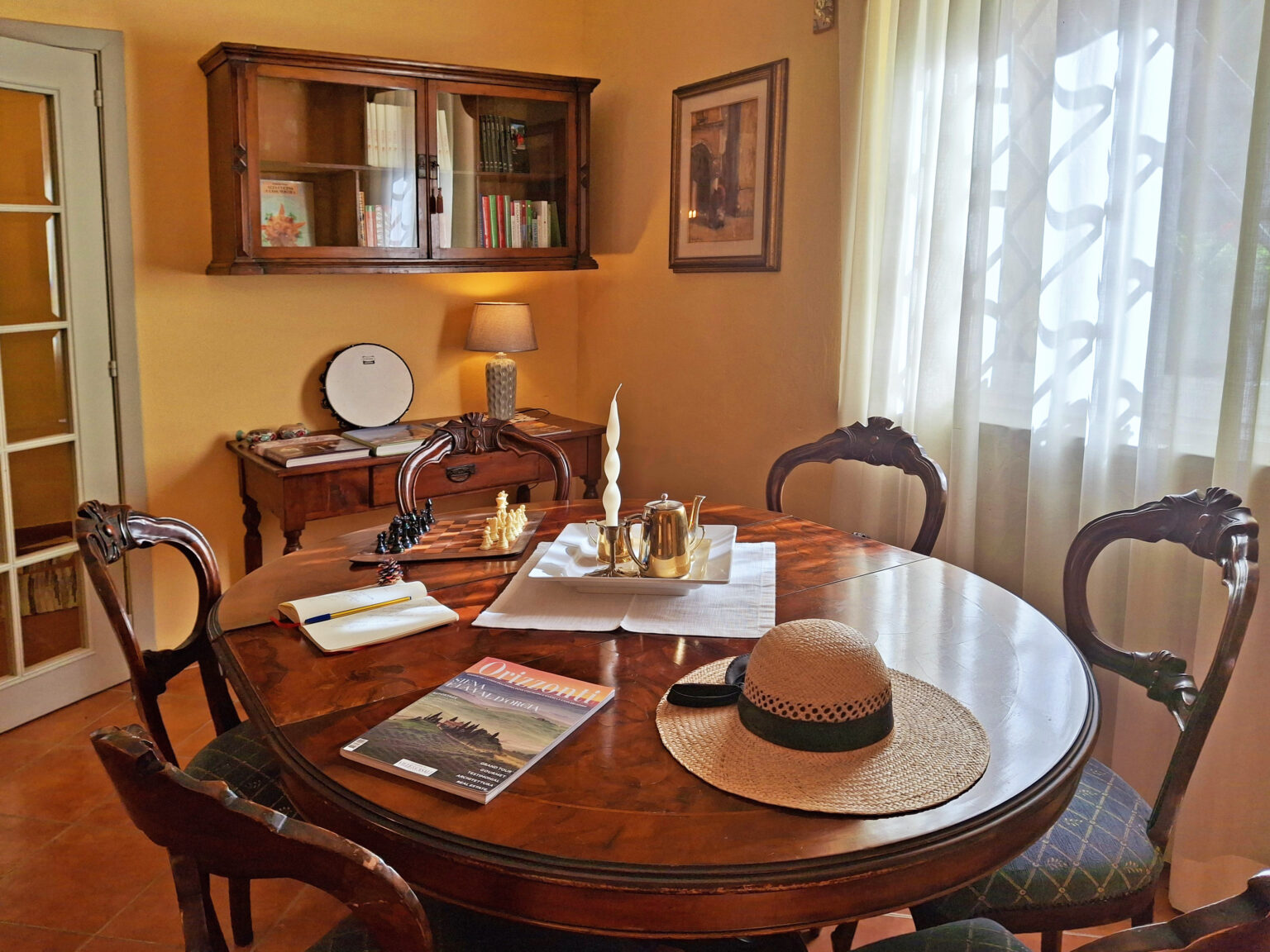 Cozy dining area featuring a round wooden table set with a hat, a magazine, and decorative items, illuminated by natural light from a window. The warm yellow walls and elegant furniture create an inviting atmosphere. | Only on SopranoVillas