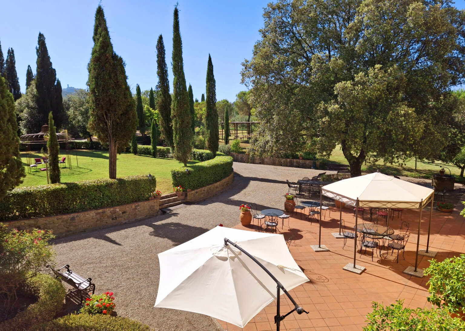 Scenic outdoor garden area featuring tall cypress trees, shaded seating under white umbrellas, and a well-maintained pathway, ideal for relaxation and gatherings. | Only on SopranoVillas