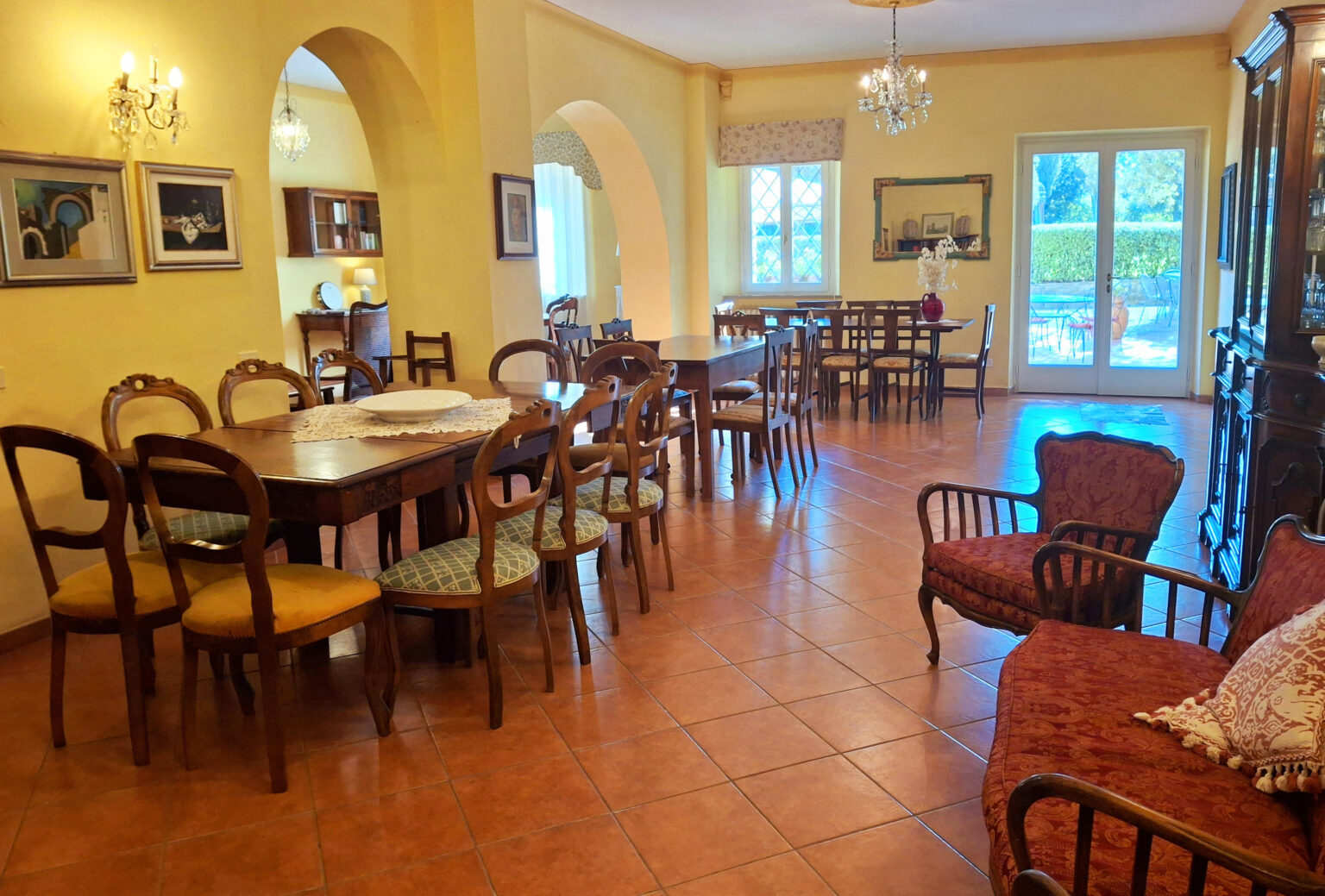Spacious dining area with wooden tables and chairs, featuring warm yellow walls and natural light from large windows, creating an inviting atmosphere for gatherings. | Only on SopranoVillas