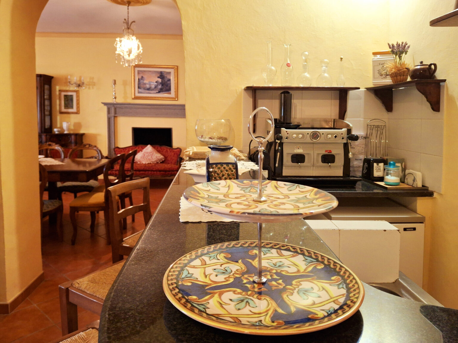 Cozy kitchen interior featuring a coffee machine, decorative plates, and a dining area in the background, with warm yellow walls and wooden furniture. | Only on SopranoVillas
