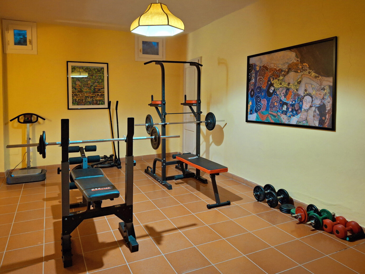 Home gym setup featuring a weight bench, barbell rack, dumbbells, and fitness equipment against a yellow wall with artwork. Ideal for strength training and fitness enthusiasts. | Only on SopranoVillas