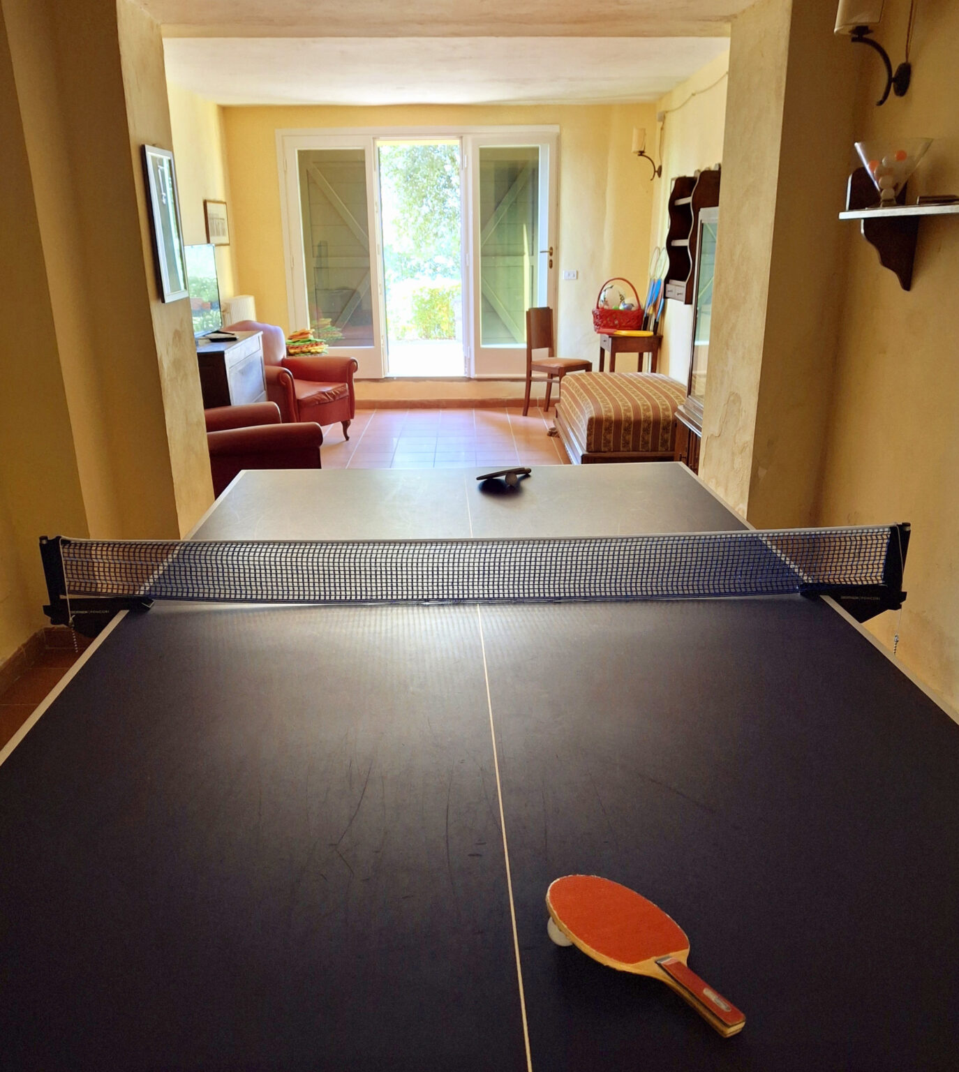 Interior view of a ping pong table with a net, set up in a cozy room featuring a sofa and chairs in the background, showcasing a recreational space for indoor sports. | Only on SopranoVillas