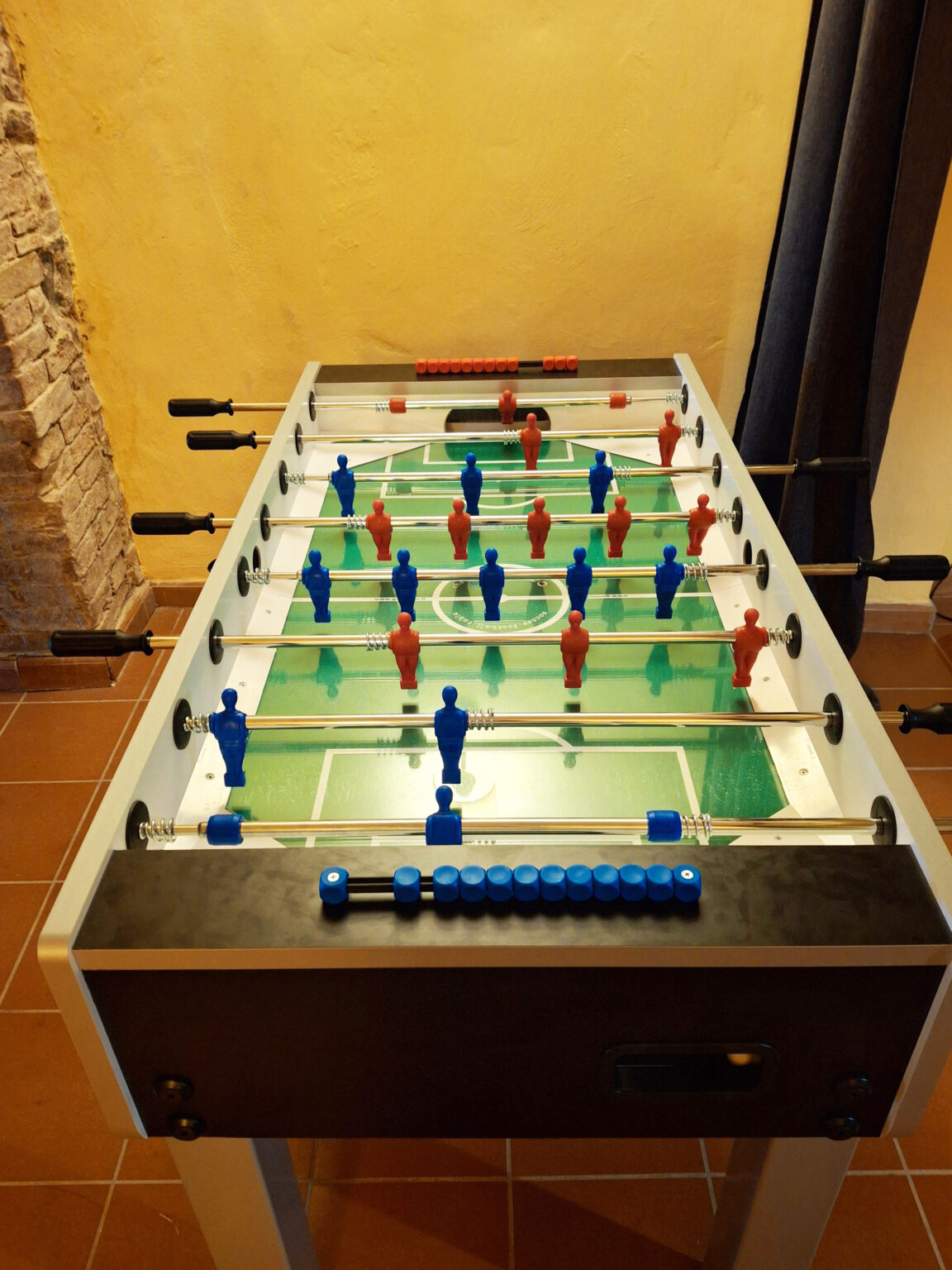 Foosball table featuring blue and red player figures arranged on a green playing field, set against a textured yellow wall. The table includes rods for player manipulation and a scoreboard on the side, ideal for indoor game rooms or recreational spaces. | Only on SopranoVillas