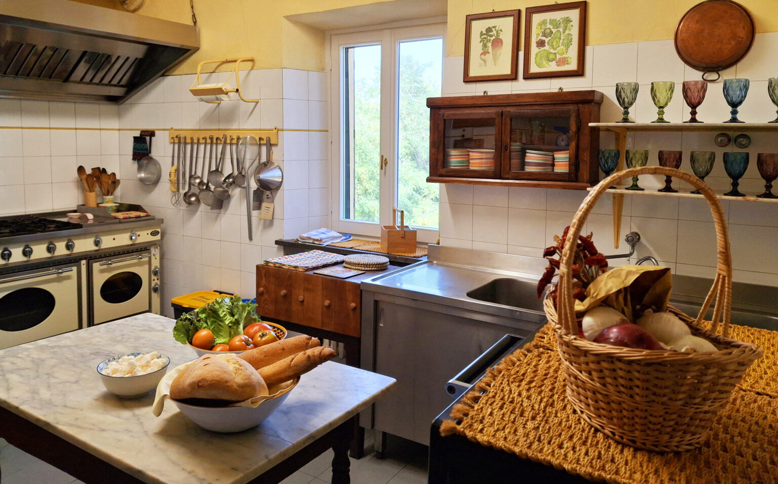 Cozy kitchen interior featuring a wooden dining table with fresh vegetables, a basket of assorted produce, and vintage kitchenware. The space is illuminated by natural light from a window, highlighting the rustic decor and functional appliances. | Only on SopranoVillas