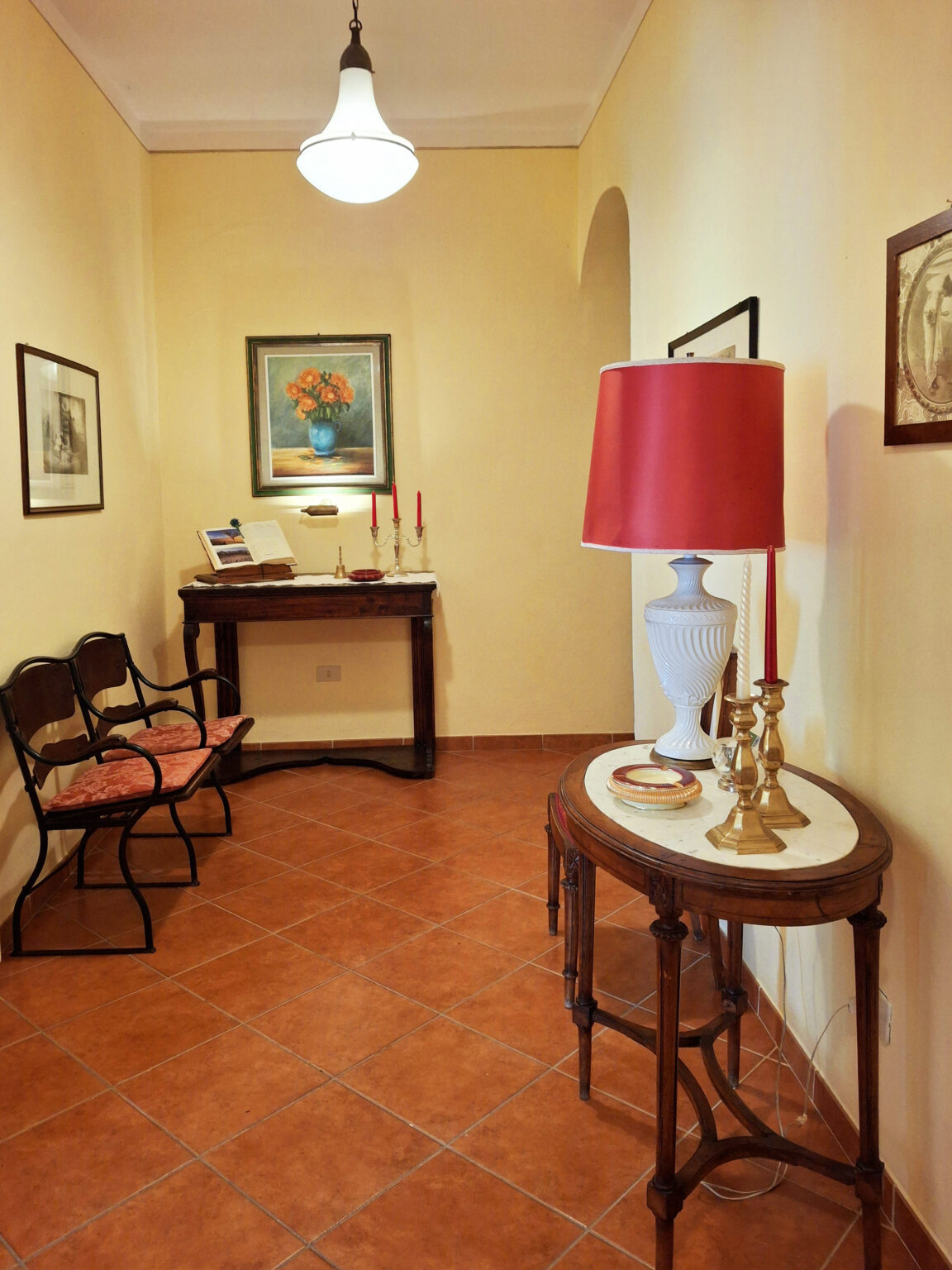 Cozy hallway interior featuring a decorative table with a white lamp, red lampshade, and vintage candle holders. Two stylish chairs with cushions and framed artwork adorn the walls, creating an inviting atmosphere. | Only on SopranoVillas