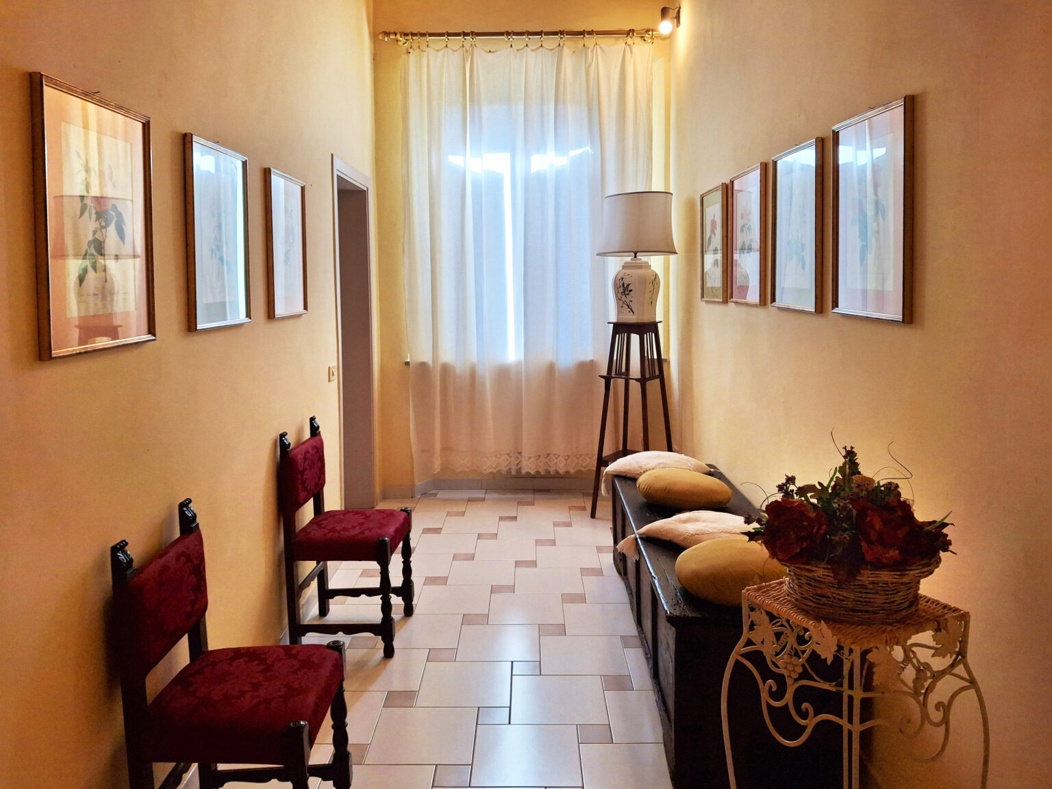 Bright hallway featuring framed botanical art, two red upholstered chairs, a decorative lamp on a wooden stand, and a bench with cushions. Soft natural light filters through sheer curtains, enhancing the warm ambiance of the space. | Only on SopranoVillas