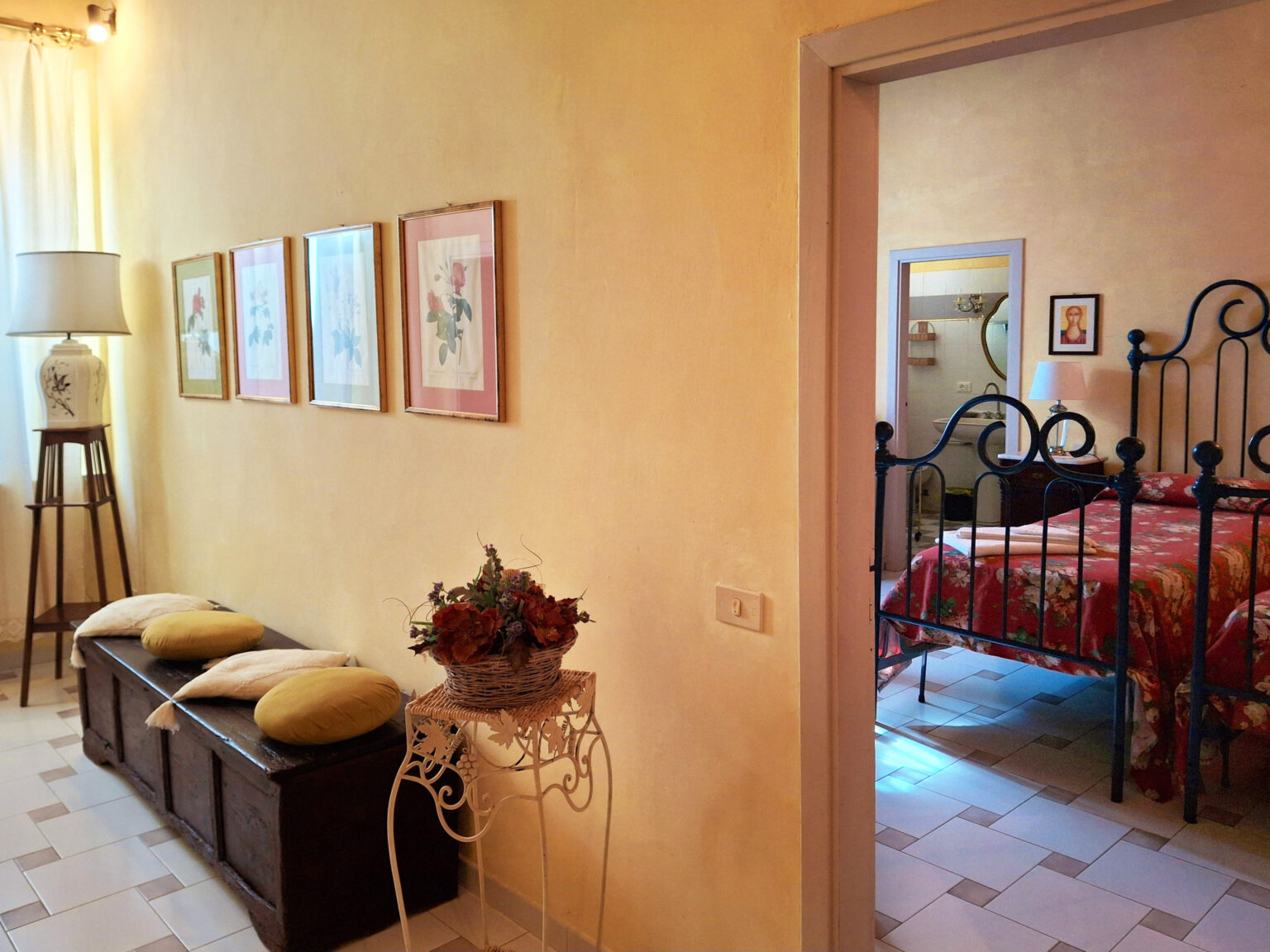 Cozy hallway featuring framed floral artwork, a vintage lamp, and a wooden bench with cushions, leading to a warmly decorated bedroom with an iron bed frame and soft lighting. | Only on SopranoVillas