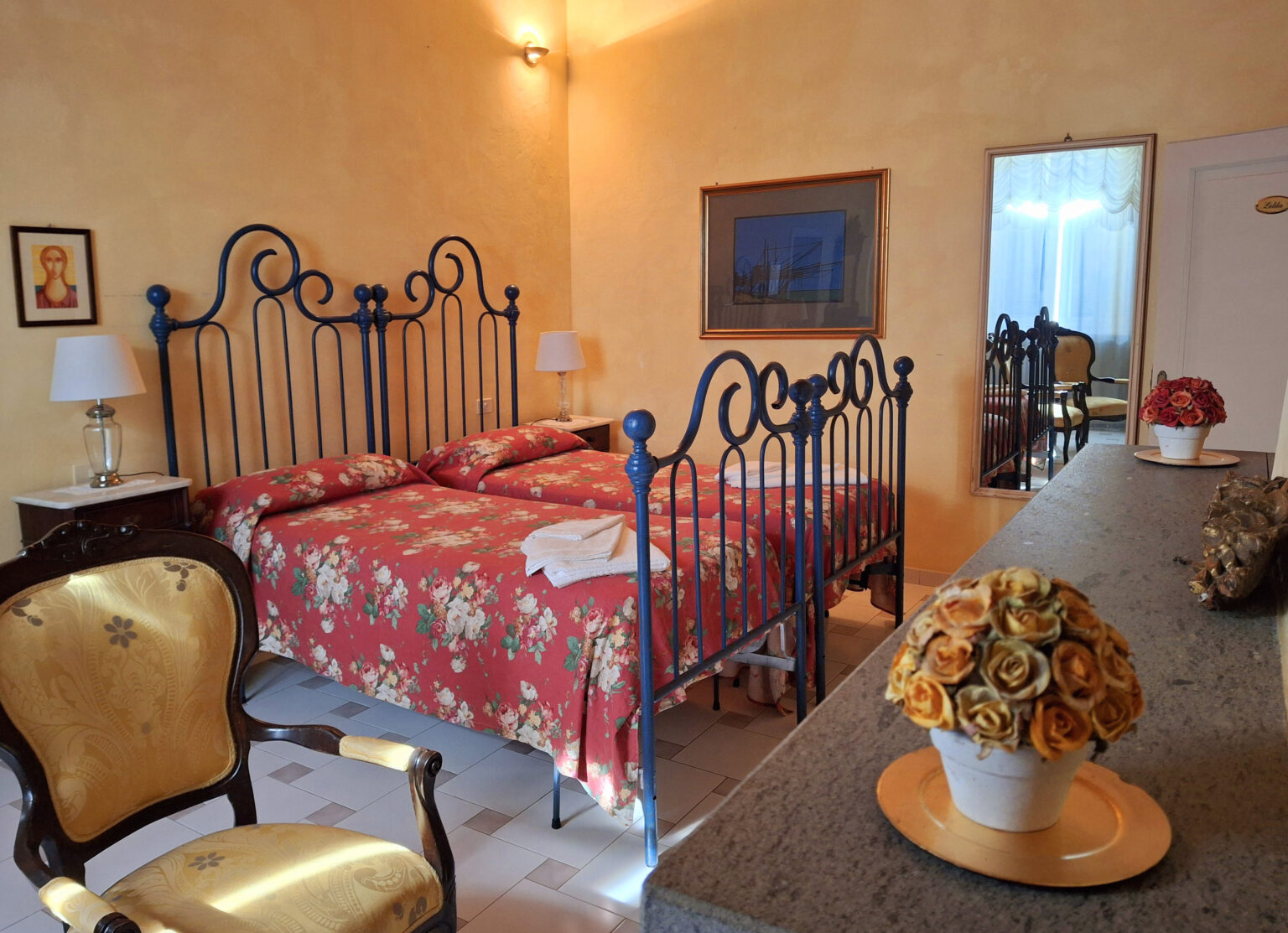 Cozy hotel room featuring a floral-patterned double bed with wrought iron headboards, elegant wooden furniture, and decorative accents, creating a warm and inviting atmosphere. | Only on SopranoVillas