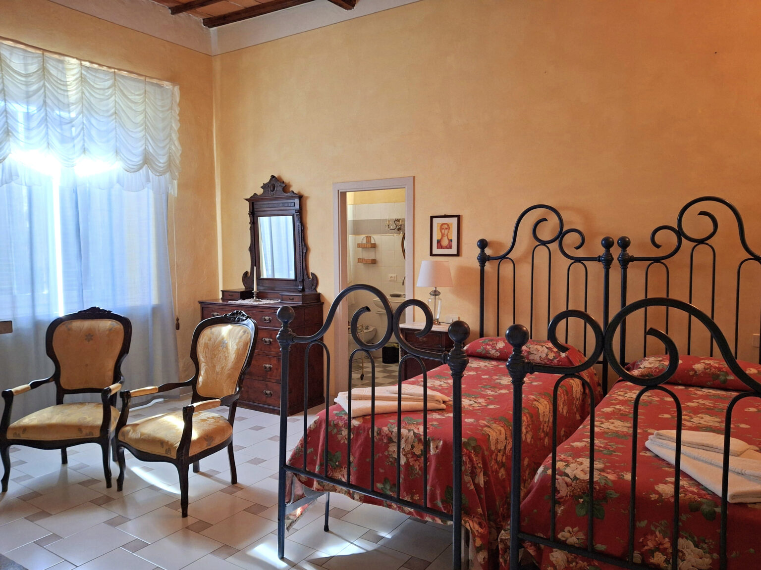 Cozy bedroom featuring two floral-patterned beds with black wrought iron frames, ornate wooden chairs, a vintage dresser, and a large window draped with sheer curtains, creating a warm and inviting atmosphere. | Only on SopranoVillas