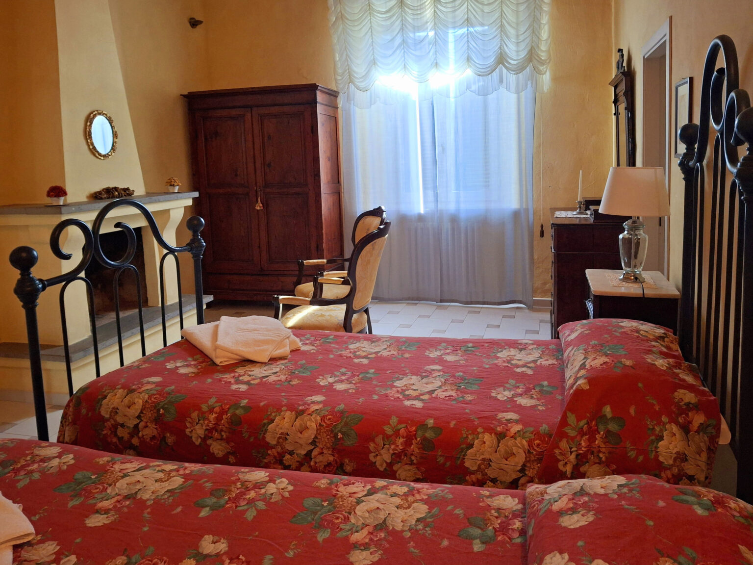 Cozy bedroom featuring two iron bed frames with floral bedding, a wooden wardrobe, an armchair, and a bright window with sheer curtains, creating a warm and inviting atmosphere. | Only on SopranoVillas