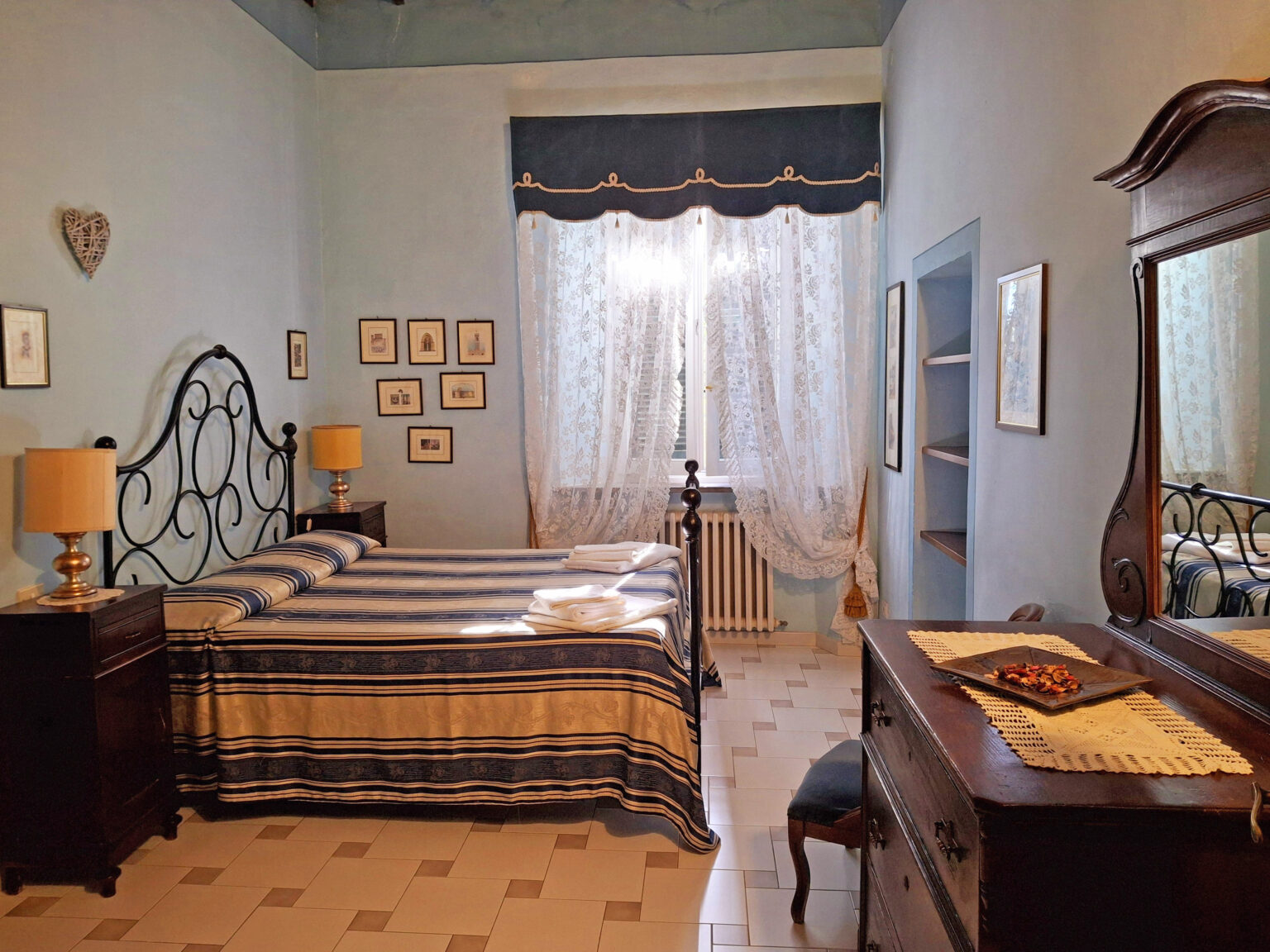 Cozy bedroom featuring a queen-sized bed with blue and white striped bedding, vintage wooden furniture including a dresser and bedside lamps, and large windows adorned with sheer curtains, creating a bright and inviting atmosphere. | Only on SopranoVillas