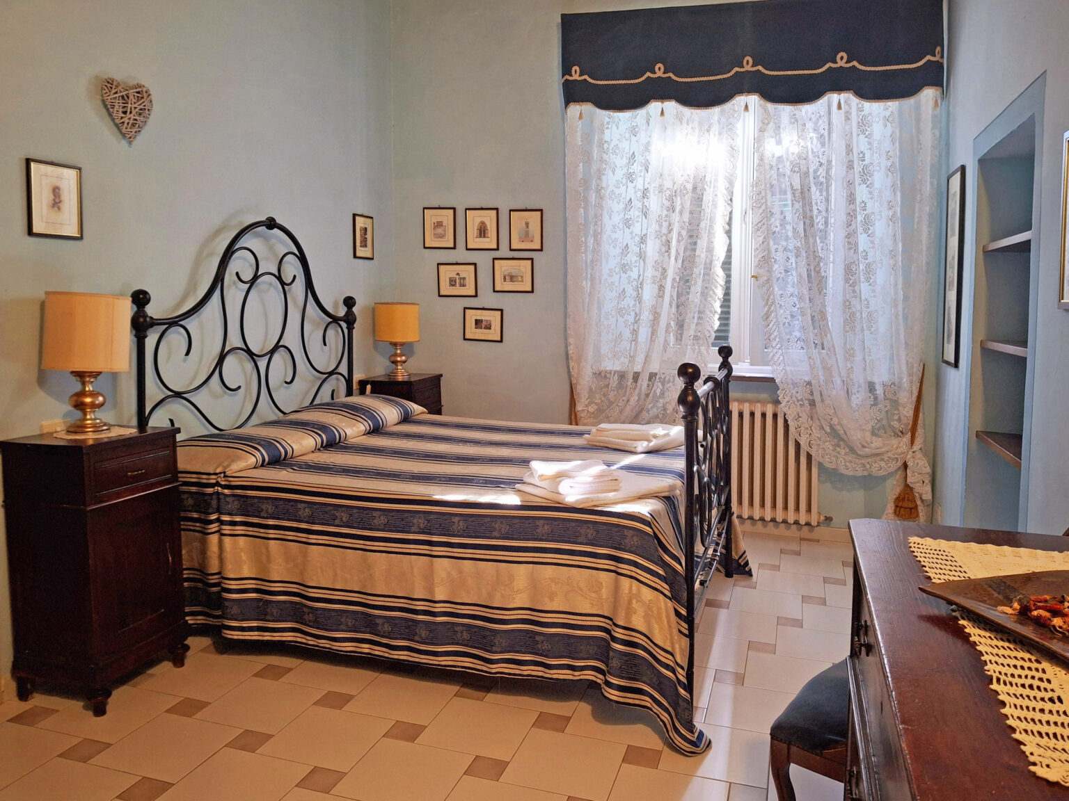 Cozy bedroom featuring a blue color scheme, a wrought iron bed with striped bedding, wooden nightstands, and framed artwork on the walls, illuminated by natural light from a large window. | Only on SopranoVillas