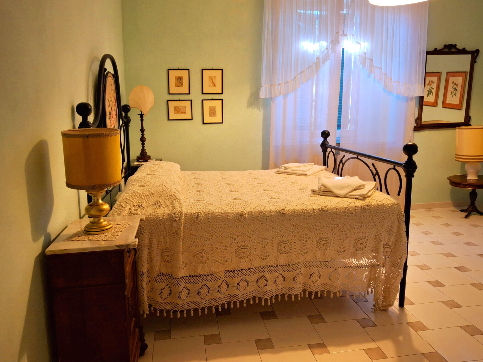Cozy bedroom featuring a wrought iron bed with a lace bedspread, vintage lamps, and framed botanical prints on light green walls. Natural light filters through sheer curtains, enhancing the inviting atmosphere. | Only on SopranoVillas