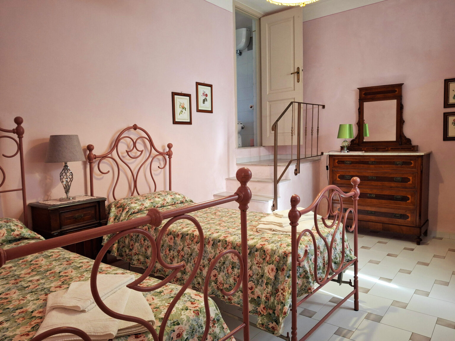 Cozy bedroom featuring two vintage iron beds with floral bedding, a wooden dresser, and soft pink walls, creating a warm and inviting atmosphere. | Only on SopranoVillas