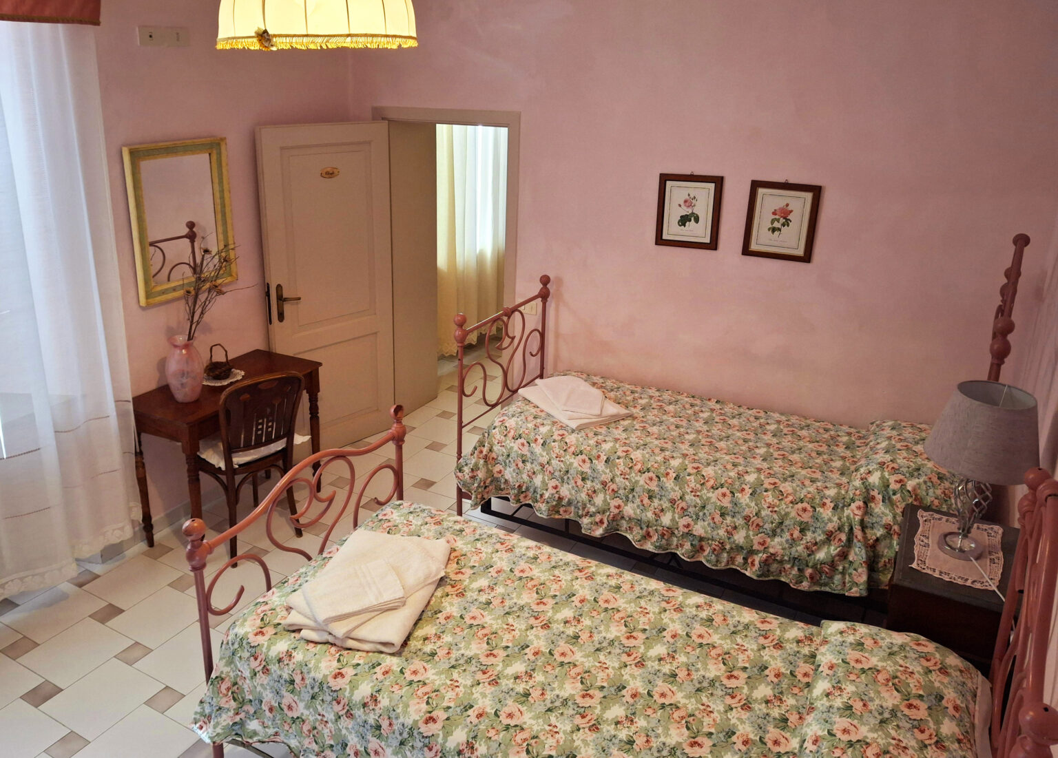 Cozy bedroom featuring two twin beds with floral bedspreads, a wooden desk and chair, and soft lighting. The walls are painted pink, complemented by decorative framed botanical prints. Natural light filters through sheer curtains, creating a warm and inviting atmosphere. | Only on SopranoVillas