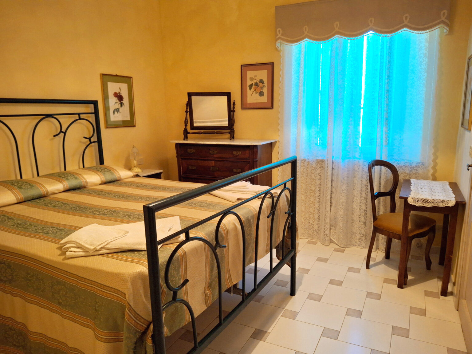 Cozy bedroom featuring a wrought iron bed with decorative bedding, a wooden dresser, and a small writing desk, illuminated by natural light from a blue-curtained window. The warm yellow walls add a welcoming ambiance. | Only on SopranoVillas