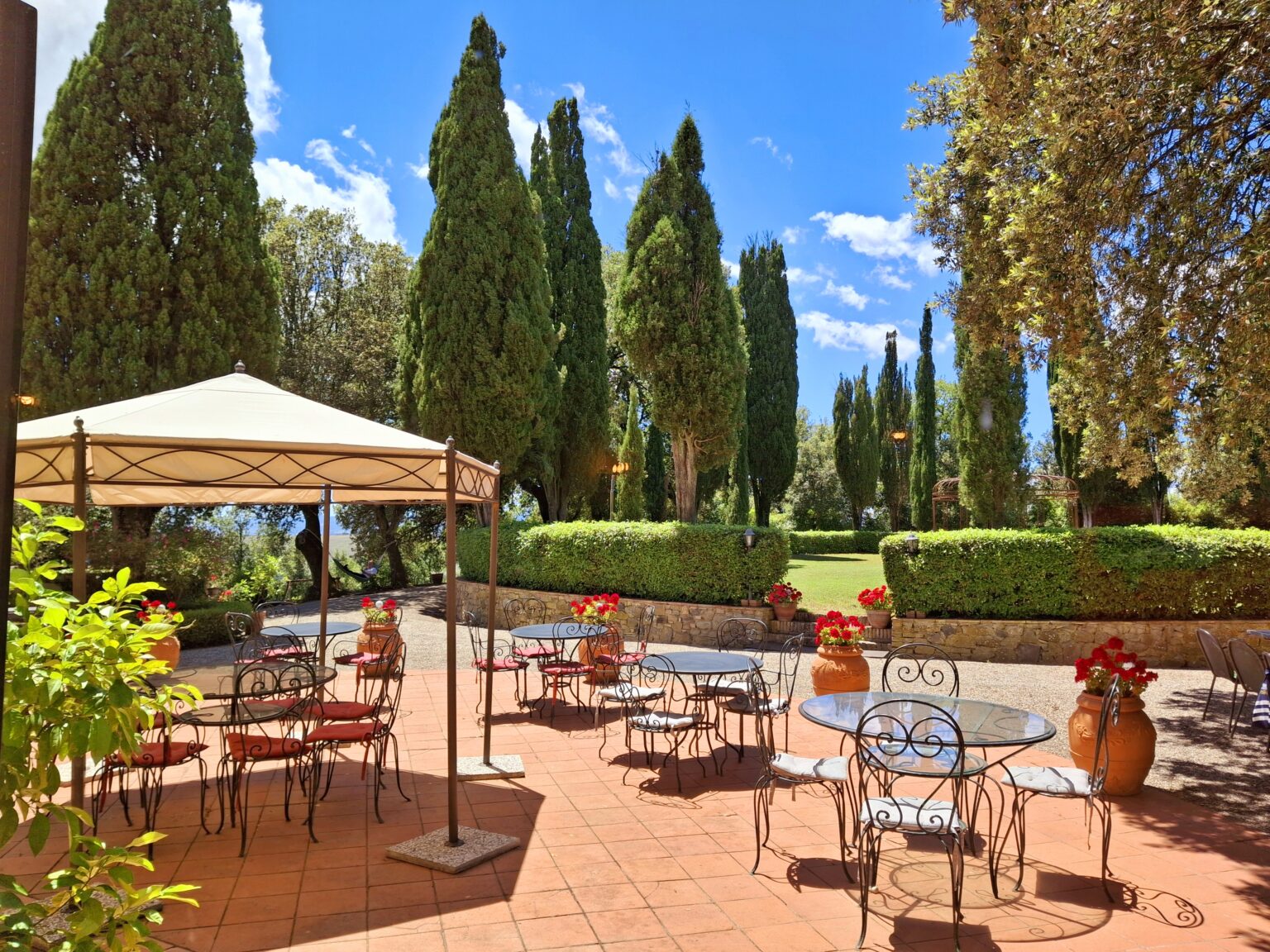 Outdoor patio area with tables and chairs under a canopy, surrounded by tall cypress trees and vibrant flowers, ideal for relaxation and dining in a scenic garden setting. | Only on SopranoVillas