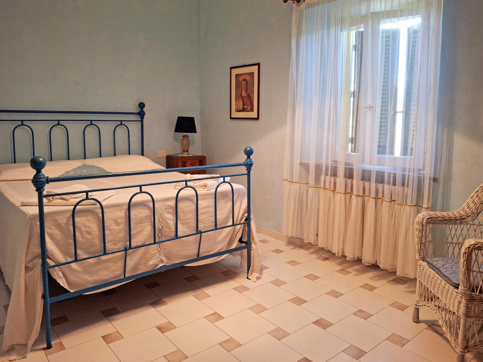 Cozy bedroom featuring a blue metal bed frame, white bedding, a bedside lamp, and a wicker chair, with soft natural light filtering through sheer curtains. | Only on SopranoVillas