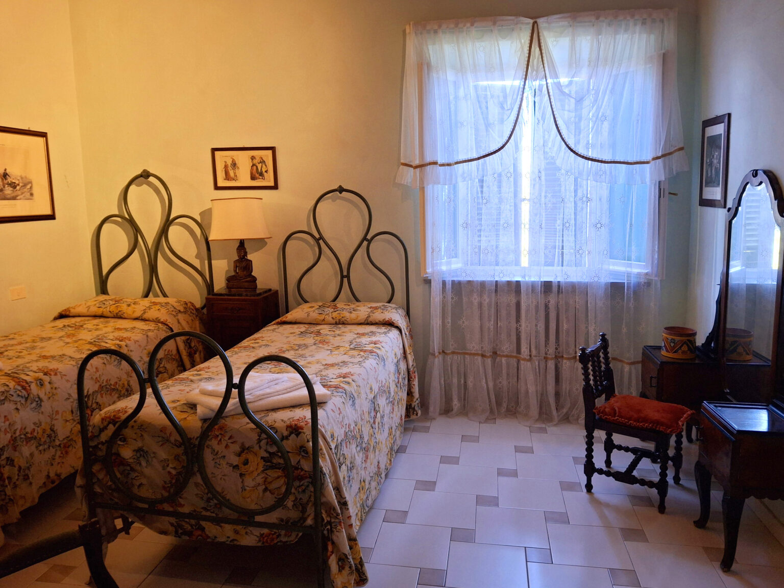 Cozy bedroom featuring two vintage iron beds with floral bedding, a wooden nightstand with a lamp, a small vanity table with a chair, and soft natural light filtering through sheer curtains. | Only on SopranoVillas