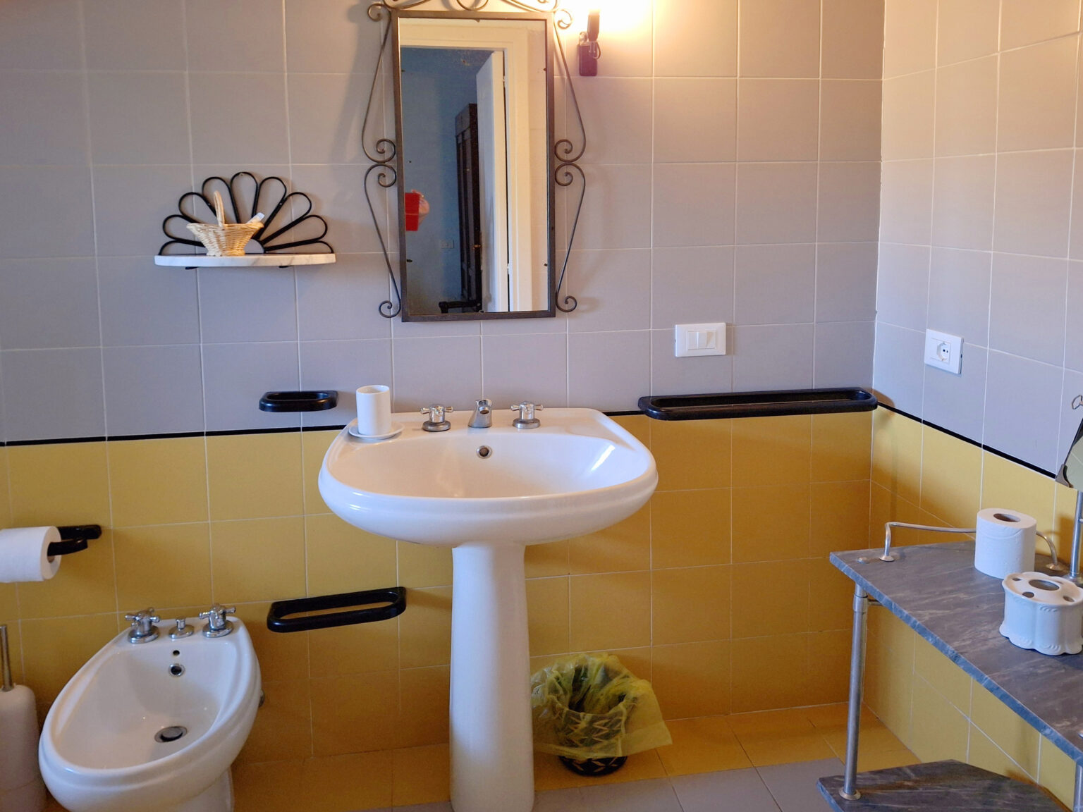 Modern bathroom interior featuring a white sink, bidet, and a large mirror. The walls are tiled in a mix of gray and yellow, complemented by decorative shelving. Essential items like toilet paper and a wastebasket are visible, creating a functional and stylish space. | Only on SopranoVillas