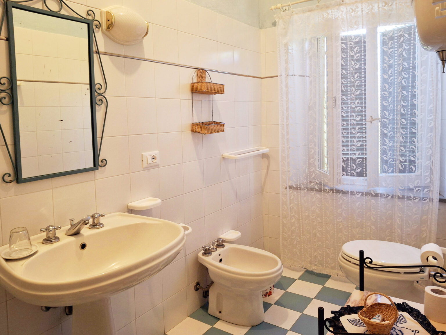 Bright and airy bathroom featuring a white sink, bidet, and toilet, complemented by a large window with sheer curtains and a decorative mirror. The tiled floor showcases a classic checkered pattern. | Only on SopranoVillas