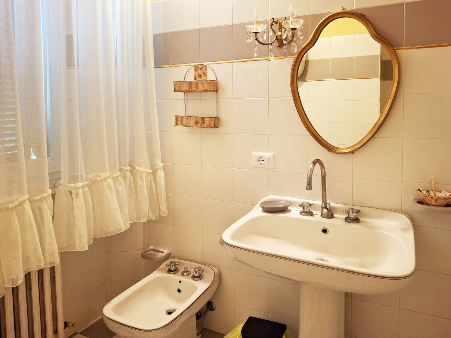Bright and airy bathroom featuring a white sink, bidet, and a round mirror framed in gold. Soft white curtains enhance the natural light, while wooden shelves add a rustic touch. The tile walls provide a clean and modern aesthetic. | Only on SopranoVillas