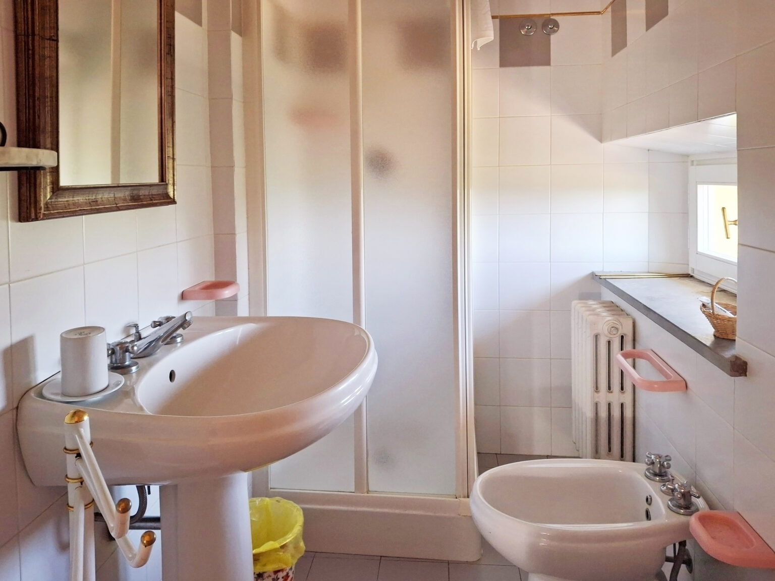 Bright and modern bathroom featuring a white sink, bidet, and a glass shower enclosure, complemented by a wooden mirror and a window that allows natural light. | Only on SopranoVillas