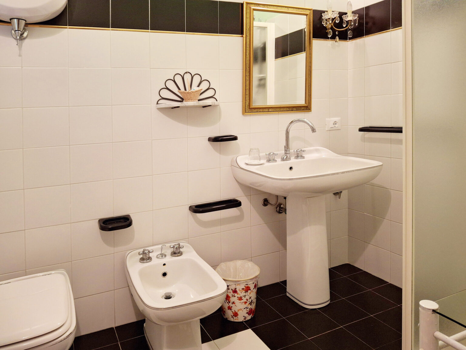 Modern bathroom featuring a white pedestal sink, bidet, and toilet against a tiled wall. The space includes a decorative mirror, wall-mounted shelves, and a stylish wastebasket, creating a clean and functional design. | Only on SopranoVillas