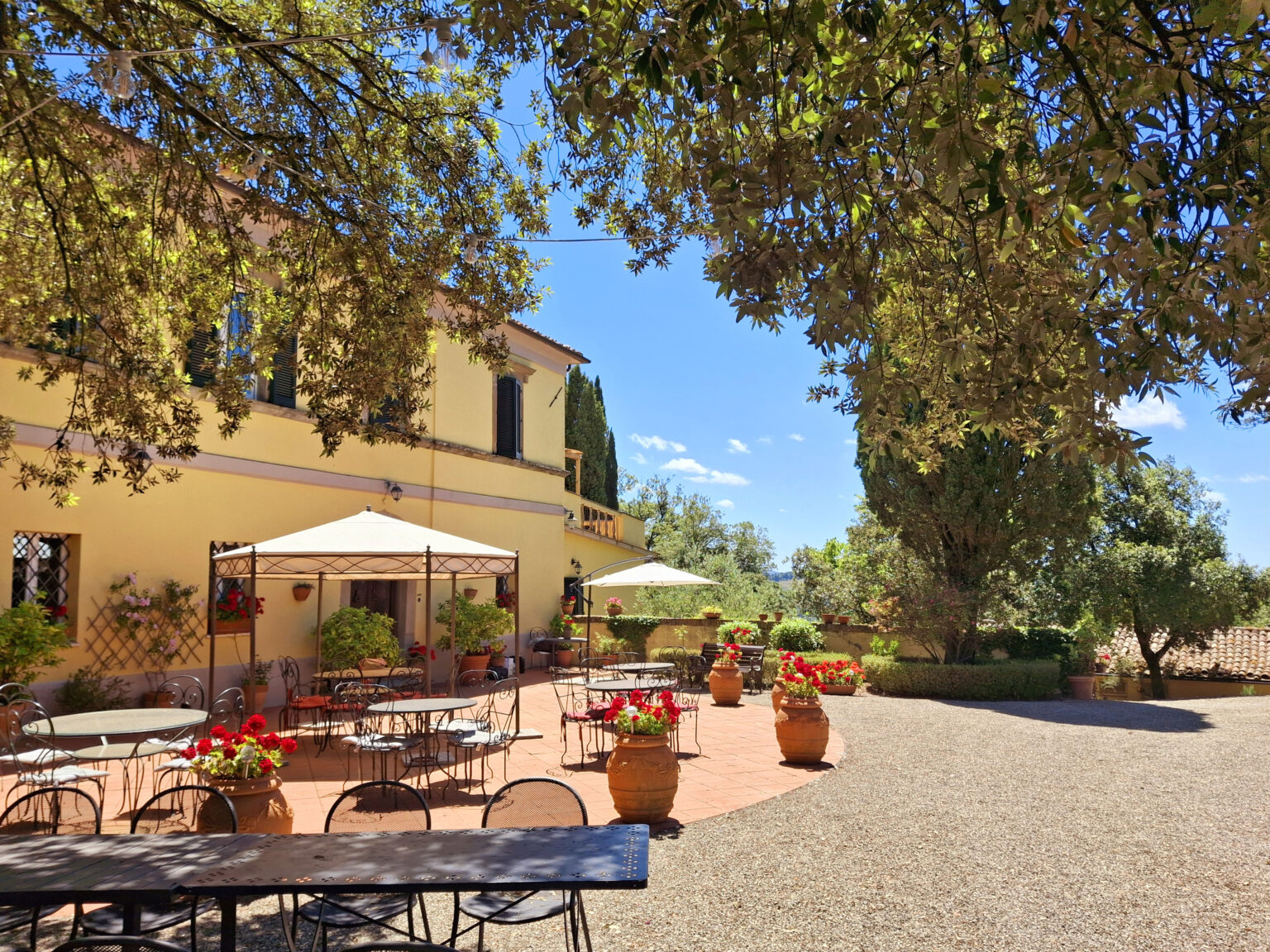 Charming outdoor dining area at a rustic Italian villa, featuring tables under umbrellas, vibrant flower pots, and lush greenery against a clear blue sky. Ideal for enjoying meals in a scenic setting. | Only on SopranoVillas