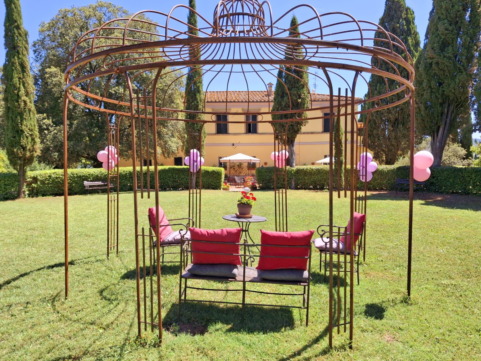 Decorative outdoor gazebo with red cushions, surrounded by green grass and trees, set in a sunny garden. Ideal for gatherings and relaxation. | Only on SopranoVillas